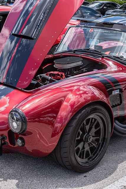 Red classic sports car with black racing stripes and hood open showing engine at outdoor car show